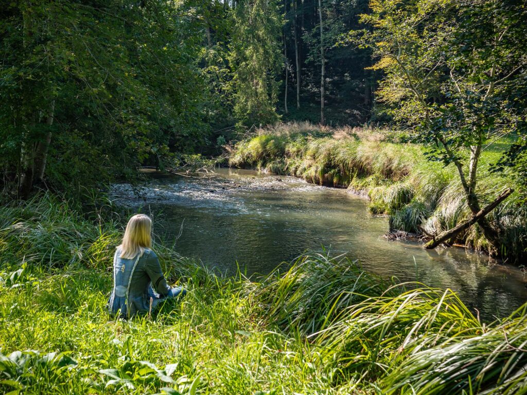 Frau sitzt meditierend an einem idyllischen Bach am Waldrand
