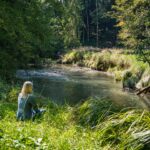 Frau sitzt meditierend an einem idyllischen Bach am Waldrand