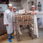 Die Metzgermeister Sichert und Braunsperger zusammen mit Dr. Peter Loreth, Leiter der Verwaltungstelle der Biosphärenregion, mit den für den Räucherprozess vorbereiteten Biosphären-Kabanossi und Biosphären-Produkt-Schild